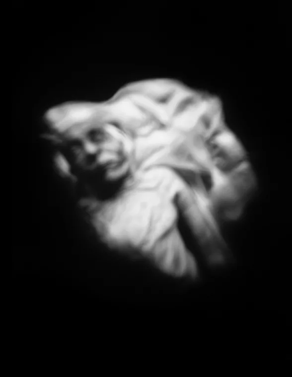 A blurry black and white photo shows a head and chest of a woman who is deceased. Her with eyes and mouth look open and the images is surrounded by deep black background. The image appears to be like a ghostly apparition. 