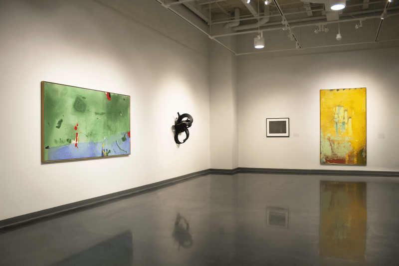 A large museum gallery with a polished concrete floor shows a group of four abstract art works, including a large green and blue horizontal piece, a black sculptural ornament, a framed work on paper and a yellow vertical piece.