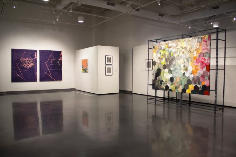 A large museum gallery with a polished concrete floor shows a group of abstract art works, including a purple-pink diptych, a small orange and yellow painting, several framed small works and a large painting on a scaffolding that is made up of cube and hexagon shapes in varying colors from pink to yellow and black, seeming a kind of large honeycomb.