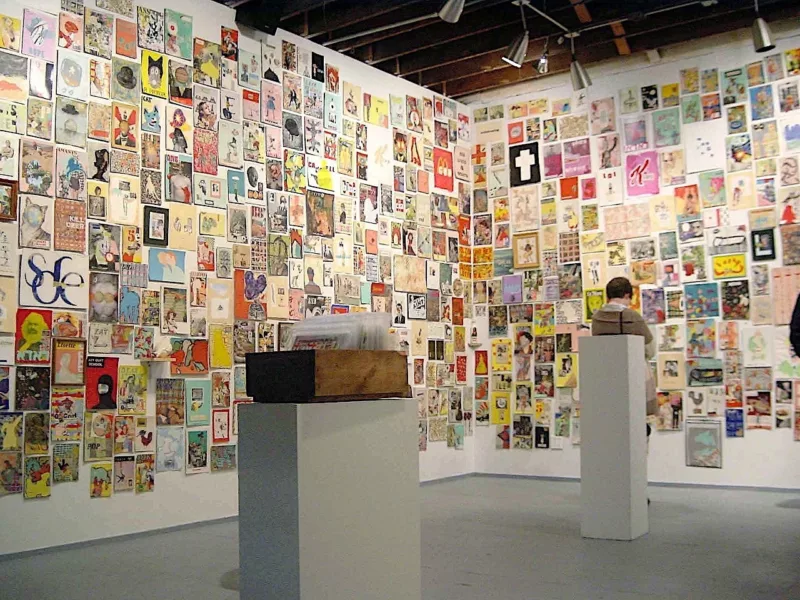 An art gallery with concrete floor and exposed wood beams in the ceiling shows two walls adjoining in a corner that are filled with colorful small art pieces hung like a wallpapering of the walls. Two pedestals have boxes filled with more small works. A gallery goer is studying the art in the background.