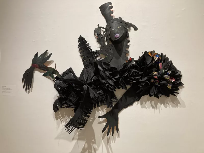 A large complex cut paper piece shows a woman’s head and upper torso and arms, perhaps dancing, perhaps cooking up a brew, her arms and torso adorned by paper pieces that look like feathers or perhaps shells, some colored bright red or golden. 
