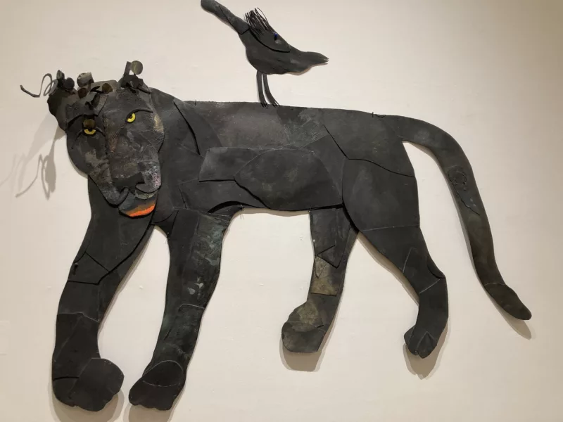 A large cut paper piece of a black panther stares at you with its intense yellow eyes and open mouth painted bright red depicts a regal animal, healthy and alert. A Black bird companion perches on its back, tail up and head out, also alert and poised.