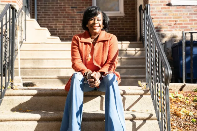 A smiling Black woman wearing blue jeans and an orange sweater sits on the stoop in front of a Philadelphia row house. She is Cherelle Parker and she is the incoming Mayor of the City of Philadelphia.