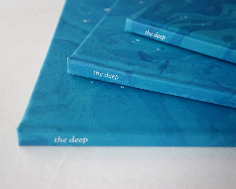 Three books, titled "the deep," whose covers are a beautiful deep aqua blue, rest on top of each other on a white surface with their spines facing towards you.