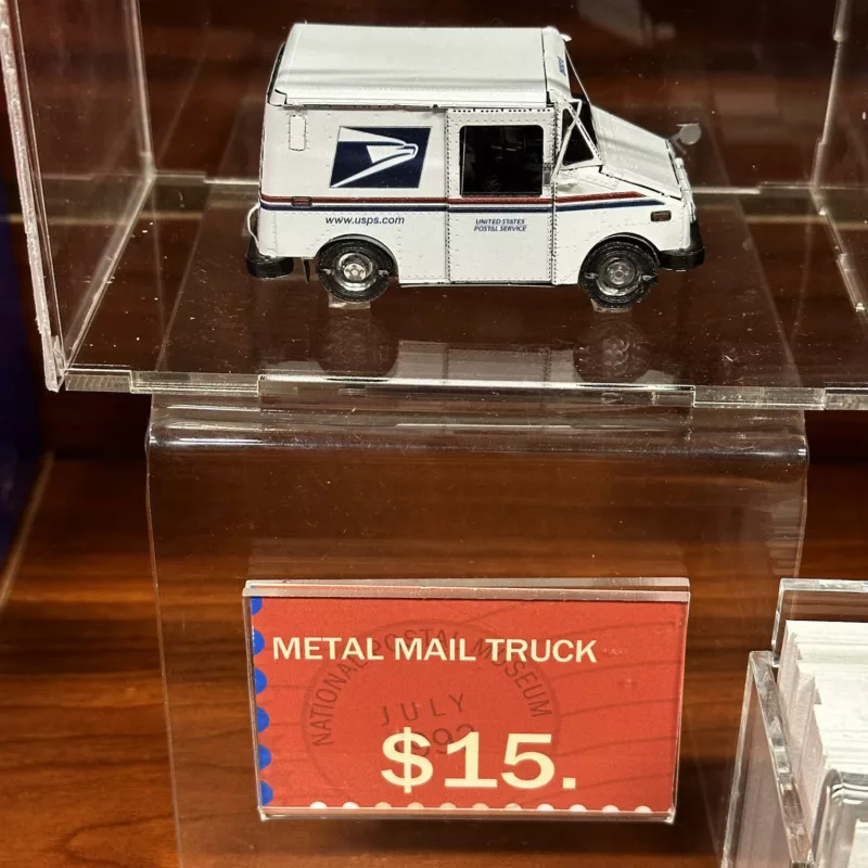 A familiar white USPS mail truck in miniature sits parked in a plastic vitrine above a sign that says, “metal mail truck” $15.
