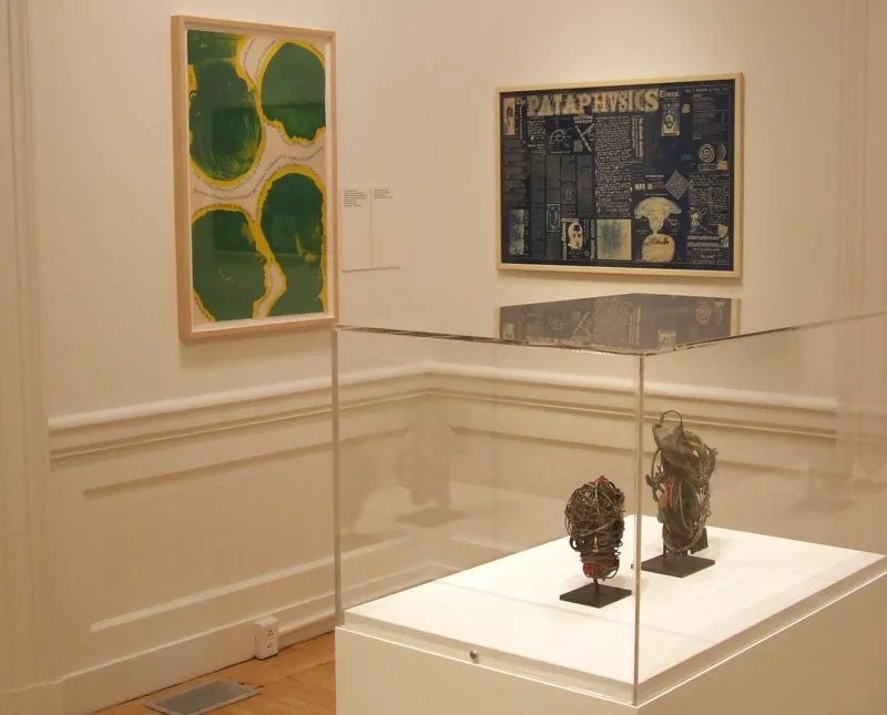 An art installation in a corner of a gallery with wainscoting and wood floors shows two framed works on the left and right walls and in a vitrine in the foreground, two small abstract wire sculptures.