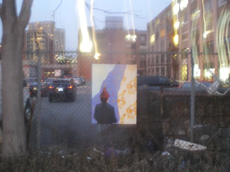 A painting hanging on a cyclone fence in front of an apartment parking lot shows a person facing away from you, wearing a black jacket and red dunce hat, and in the background a repeat pattern of clown faces partly covered by a violet shadow or road of red stars and yellow crescent moons that leads into the far distance.