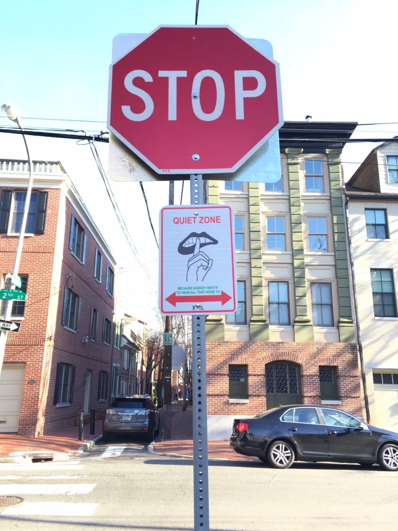 A Stop sign seems to tower over a street with row houses, directing motorists to stop, while another less familiar sign underneath shows an open mouth with a hand in a “shoosh” gesture with the label “Quiet Zone” on top of the image and below it the words “Because nobody likes to hear all that noise Yo.” With a red arrow going left and right under the sentence. 