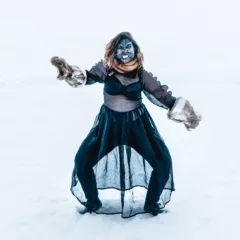 A woman stands in the snow, in a fierce pose, legs apart, knees bent, hands covered with animal fur mittens that suggest animal open mouths; her face is painted blue with a red line down the middle of the face and white lines radiating from the center outward and under the chin, and the mouth is open with the teeth bared as if growling. The woman is clothed fashionably, with a ruffled see-through dress revealing a black lace bra and leggings.