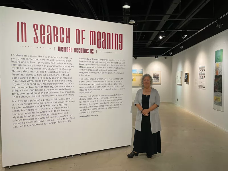 n an art gallery in a museum, a woman with curly grey hair stands smiling in front of a wall-sized sign announcing her exhibit, “In Search of Meaning: Memory Becomes Us.” She is wearing a long black dress and a grey sweater with a necklace around her neck.
