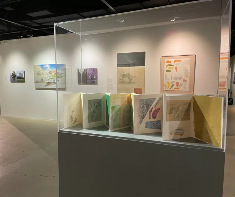  gallery is installed with paintings on the wall and in the foreground a large pedestal with a plexiglas vitrine on top holds an accordion folded book made by the artist.