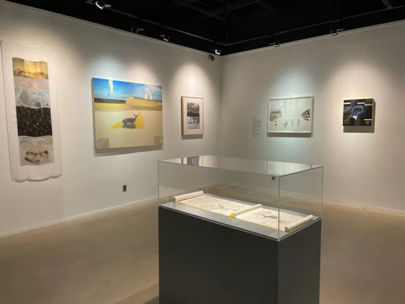 A gallery is installed with paintings and works on paper and in the foreground is a dark pedestal with a plexiglas case on top and inside the case is an open scroll with a drawing on it.