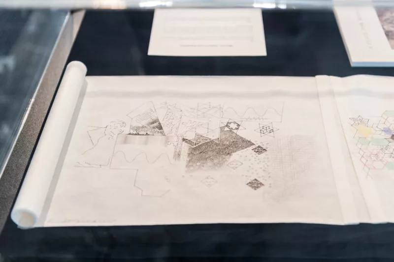 A patterned ink drawing on a rice paper scroll sits open on a black background in a glass vitrine. 
