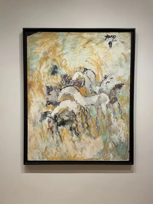 A painting in a black frame shows a herd of white and some smaller black horses painted in a stylized manner as if they are melting into a background of swirling colors, ochre, mint green and white. 