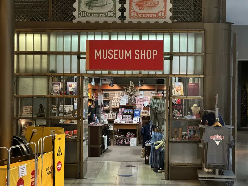 A museum shop is labeled “MUSEUM SHOP” with a red sign with white letters, and above the sign are blown up replicas of a 6 cent and a 16 cent postage stamp. The shop is the Postal Museum Shop. 