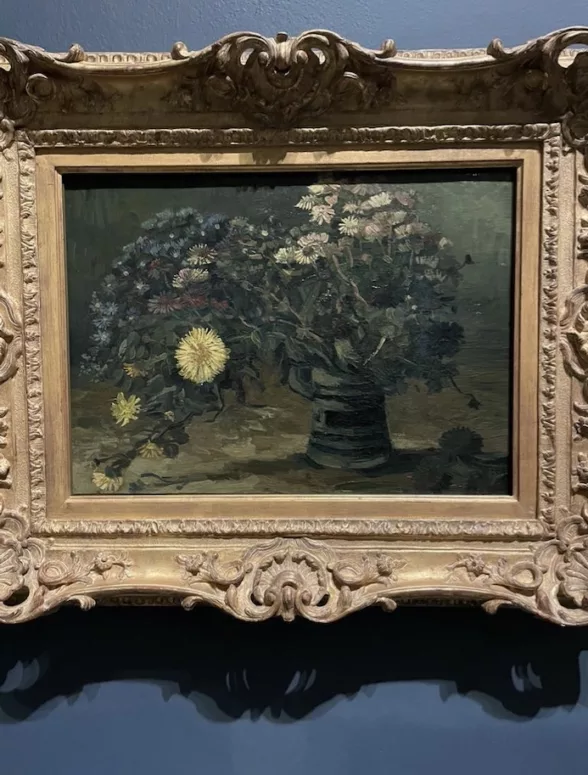 A lush bouquet of flowers sits on a table in a room with dark walls, with several flowers seeming to tip out of the vase and onto the table and others sticking straight upright.