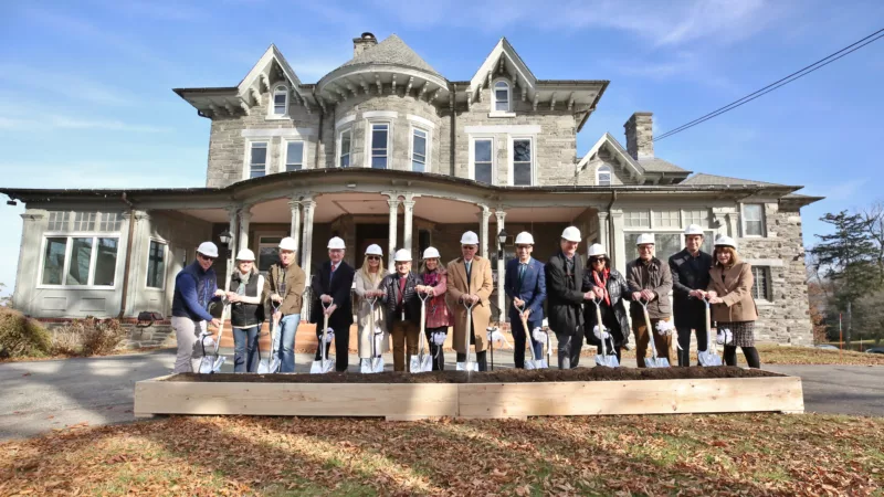 Fourteen men and women wearing white hard hats and holding shovels pose for the camera smiling in front of a large, Victorian-era stone home that will be the home of Woodmere Art Museum’s Frances M. Maguire Hall for Art and Education. The transformation of the home to the art center will be complete in 2025.