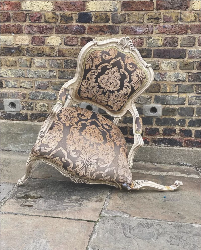 A once-ornate, now broken Versailles-looking arm chair perches forlornly on a stone pathway in front of an ancient brick wall, like a celebrity posing for a photo when they’re past their prime.