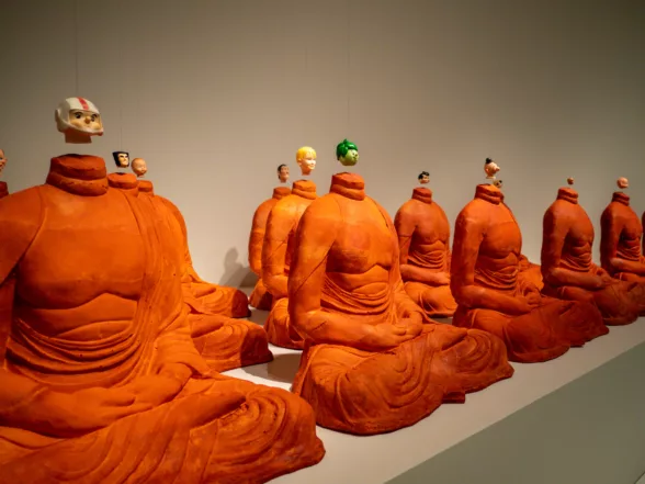 Headless, Michael JOO(마이클 주),2000 Urethane foam, vinyl plastic, styrene plastic, stainless steel wire, neodymium magnets, photo by Ryan deRoche, - 16 Buddha shaped bodies with their heads replaced with doll heads from popular comics and cartoons