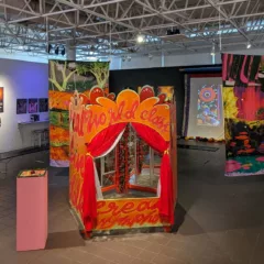 An art gallery with low lighting shows a colorful, puppet-show like cubicle and other hanging colorful works.