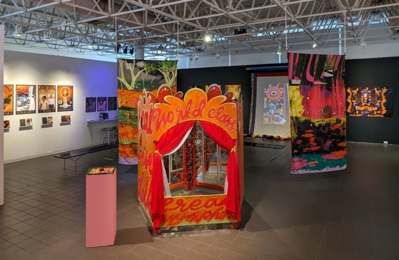 An art gallery with low lighting shows a colorful, puppet-show like cubicle and other hanging colorful works. 