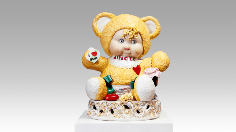 A ceramic sculpture by Jeff Koons of a child-like teddy bear embellished with hearts and sentiments of love. The sculpture sits atop an ornate gold leaf-enhanced ceramic pedestal.