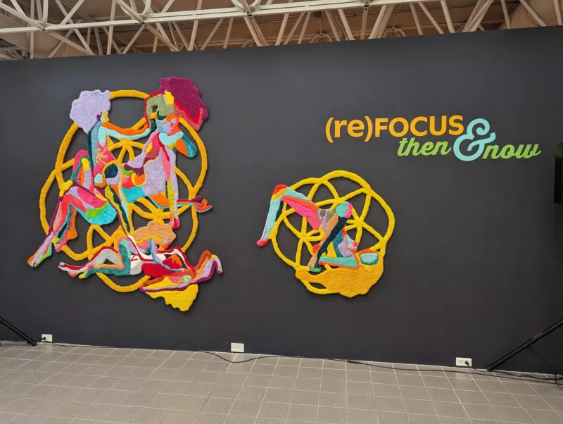 A black-painted wall announces an exhibition with words and two strong and colorful images. The words are “(re)FOCUS: Then and Now”.