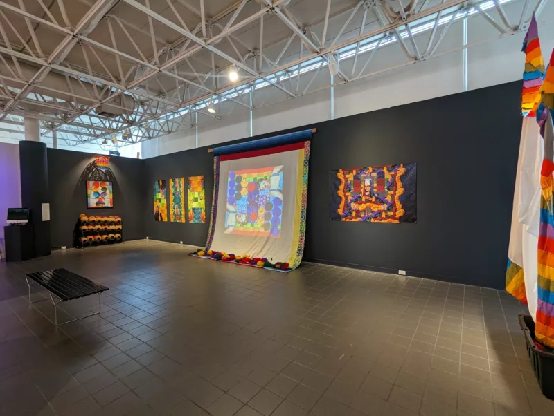 A gallery with a complex ceiling with criss-crossed metal beams on which track lights hang is installed with one wall painted black onto which a number of colorful images in oranges, blues, yellows and reds.