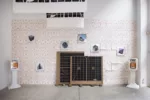 A gallery installation shows in a high-ceilinged space with a concrete floor shows a wall papered by repeat-patterned sheets of white paper with pale pink patterns on them and other objects in the space.