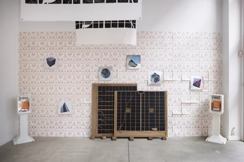A gallery installation shows in a high-ceilinged space with a concrete floor shows a wall papered by repeat-patterned sheets of white paper with pale pink patterns on them and other objects in the space.