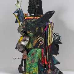 A freestanding totemic sculpture made of painted and cut paper and assembled into the shape of a small blocky human, whose Black head sits in profile on top.