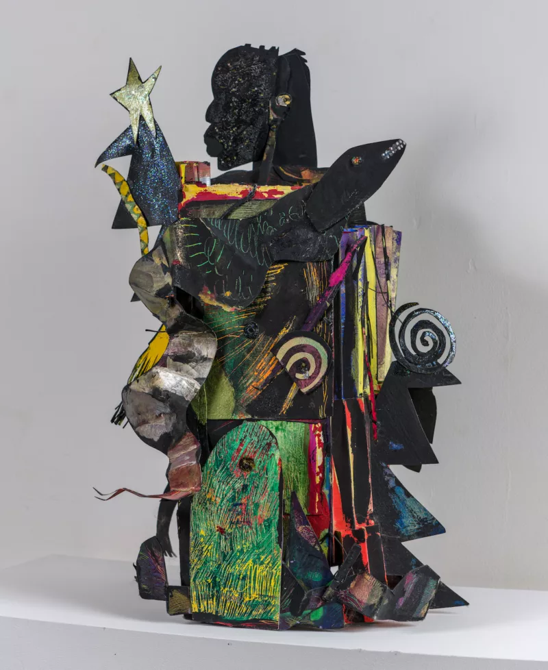 A freestanding totemic sculpture made of painted and cut paper and assembled into the shape of a small blocky human, whose Black head sits in profile on top.