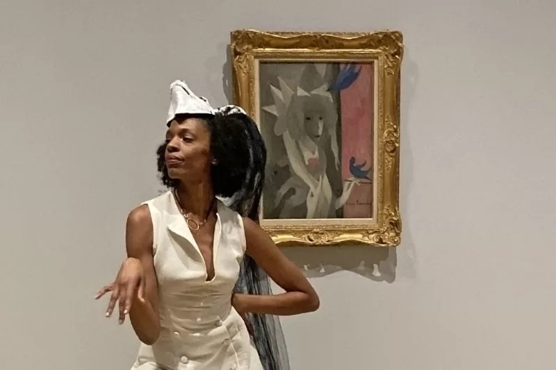 A Black woman dancer performs in front of a painting in a gold frame that represents a stylized image of a woman, interpreting the painting in her body language and costume of stylish white sleeveless dress and a white animal mask on her head.