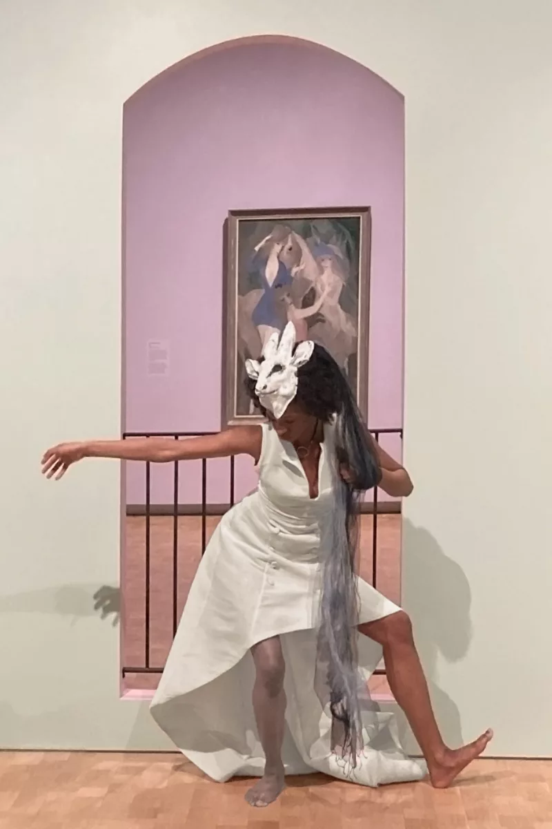 A Black woman dancer performs in front of a painting seen through a portal in a wall blocked off by a hand railing. The dancer is interpreting the painting and the railing with her body language, and her costume of stylish white sleeveless dress and a white animal mask on her head echoes the style of the work.
