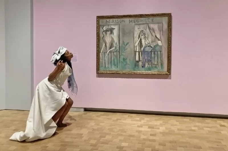 A Black woman dancer performs in front of a painting in a gold frame that represents two stylish women looking out from side by side balconies in a building called “Maison Meubleé”. The dancer is reacting to the painting in her body language, and her costume mimics the stylish garb of the women in the painting