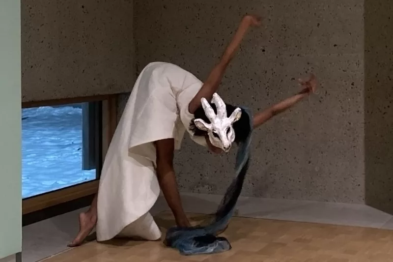 A Black woman dancer performs, interpreting the art work of Marie Laurencin in her body language and costume of stylish white sleeveless dress and a white animal mask on her head.