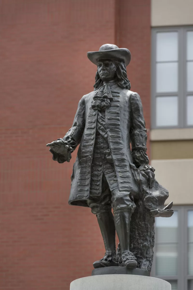 A bronze statue of the colonial founding father of Philadelphia and Pennsylvania, William Penn, wearing a broad brim hat, a fancy shirt with ruffles, a luxurious knee-length coat and breeches and boots with fancy buckles stands atop a pedestal in front of a red brick building in Welcome Park in Philadelphia.