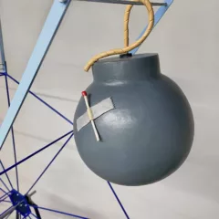 A detailed look at a cartoonish, 3D sculptural bomb with a matchstick taped to its side that is part of a larger work of a ferris wheel onto which objects like the bomb are attached and spin slowly up and down mimicking a real ferris wheel.