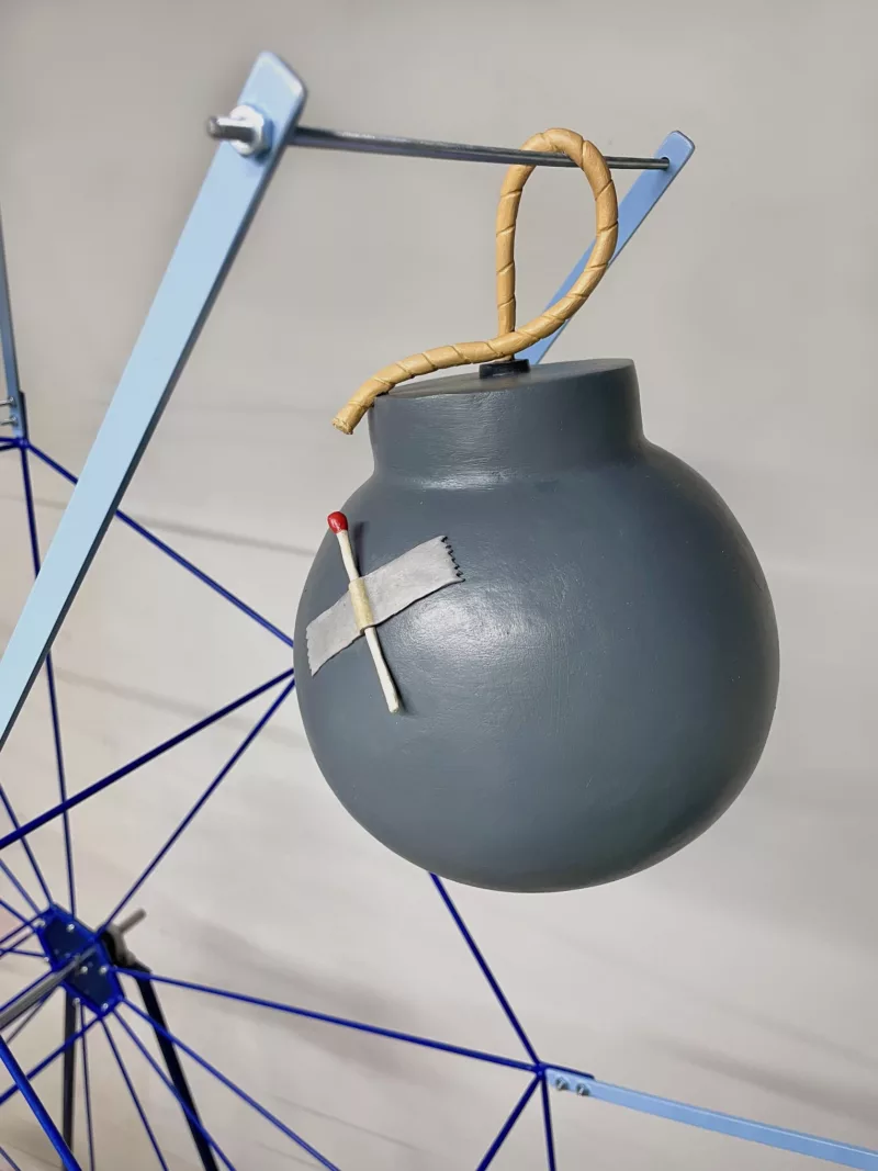 A detailed look at a cartoonish, 3D sculptural bomb with a matchstick taped to its side that is part of a larger work of a ferris wheel onto which objects like the bomb are attached and spin slowly up and down mimicking a real ferris wheel.