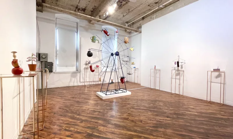 A gallery with two windows, an exposed-beam ceiling and wood floor shows a sculptural ferris wheel in the middle of the room with objects (a fire extinguisher, watering can, cartoon bomb, piece of Swiss cheese, red balloon) taking a ride up, down and around. 