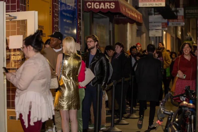 A group of mostly young people is lined up on a sidewalk waiting to be let in to an art opening at a brand new gallery building now owned by beloved gallerists who got priced out of their former space.