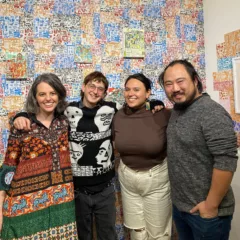 Four people with wide smiles gather together for the camera in front of a wall of colorful art. The four include the gallery owners on the ends and their two assistants in the middle.