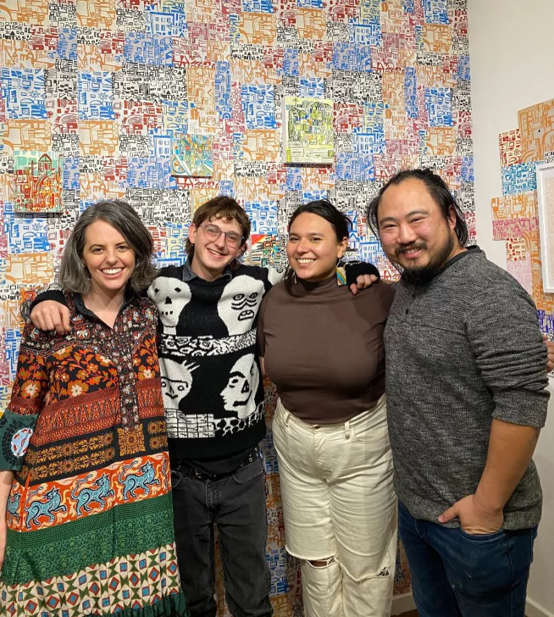 Four people with wide smiles gather together for the camera in front of a wall of colorful art. The four include the gallery owners on the ends and their two assistants in the middle.