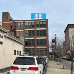 An urban street scene in Philadelphia shows cars parked in a parking space along a sidewalk and in the background, a large red brick building rises 4 stories and is topped by a billboard with a cartoon image of two robots in a deep cartoon blue background.