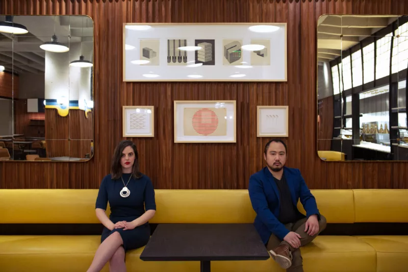 Two serious young gallerists sit on a mid-Century modern mustard-colored bench separated by a dark table in front of them. They are successful art consultants and gallery owners whose sense of play is part of their success.