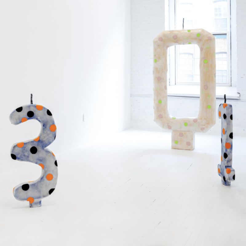 A gallery space bright lit from a large window with no curtains or shade and on the floor are three sculptures that stand up and are painted with colorful polka dots that reference parties and birthday candles in a cake — on this scale, a cake for a giant.