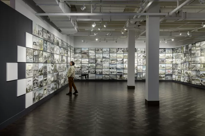 A person in a large open gallery looks up at images in a grid of mages that wrap around three walls. The floor is dark and tiled and the ceiling is open with track lighting and pipes showing.