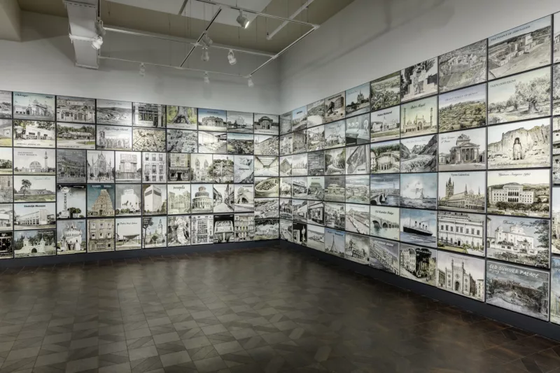 A person in a large open gallery looks up at images in a grid of mages that wrap around three walls. The floor is dark and tiled and the ceiling is open with track lighting and pipes showing.