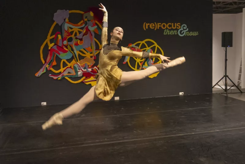 A female ballet dancer soars through the air her legs extended, toes pointed, arms in an elegant “L” surround her head; she looks exultant and in command and her costume is golden and gives her a regal look.
