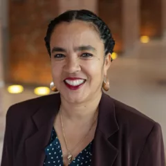 A woman wearing a big, welcoming smile looks up at the camera. She has black hair pulled back in braids, brown skin, beautiful white teeth, red lipsticked lips and is wearing hoop earrings and a wine-colored, business jacket and dark-blue, patterned blouse.